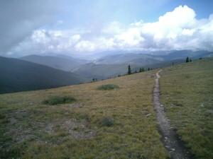 A winding dirt path leads through grassy terrain, set against a backdrop of rolling mountains and a cloudy sky. The landscape is open, showcasing the natural beauty of the highlands. Colorado Trail: Kenosha Pass To Breckenridge mountain bike trail.