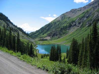 Scenic mountain landscape featuring a clear blue lake surrounded by lush green hills and evergreen trees under a bright blue sky with a few clouds. A gravel road runs alongside the lake. Trail 401 mountain bike trail.