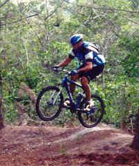 A mountain biker in mid-air, performing a jump on a dirt trail surrounded by lush green trees. The rider is wearing a blue helmet and a black and blue outfit, showcasing an adventurous moment in off-road cycling. Amelia Earhart Park mountain bike trail.