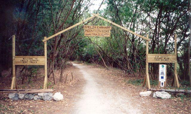 Alt text: A wooden archway marking the entrance to the Amelia Earhart Mountain Bike Trails, surrounded by greenery. Signs on either side provide trail information and welcome visitors. A dirt path leads into the wooded area. Amelia Earhart Park mountain bike trail.