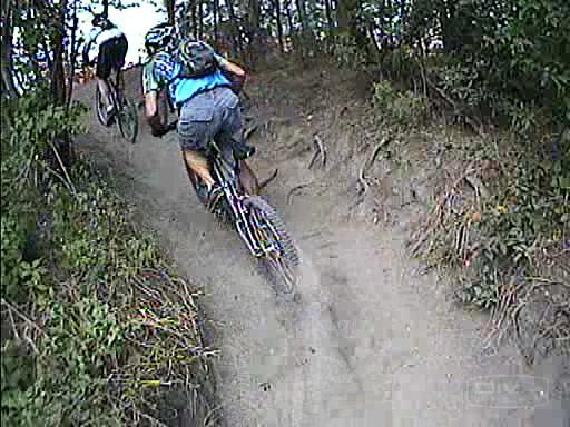 Two mountain bikers ascend a dusty trail through a wooded area, surrounded by greenery and dirt. One rider is in the foreground, wearing a blue shirt and gray shorts, with a backpack and helmet. The other biker is slightly ahead, partially visible, as they navigate the challenging terrain. Amelia Earhart Park mountain bike trail.