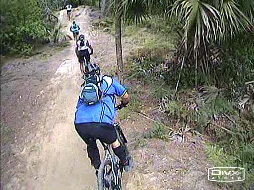 A group of mountain bikers riding along a dirt trail surrounded by greenery and trees. The lead biker is wearing a blue shirt and a backpack, with others following behind on the path. Amelia Earhart Park mountain bike trail.