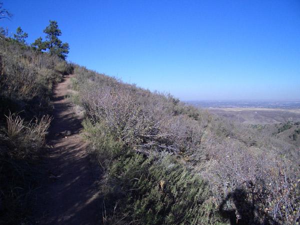 A winding dirt path through brush on a hillside with clear blue skies in the background, overlooking a valley below. Chimney Gulch mountain bike trail.