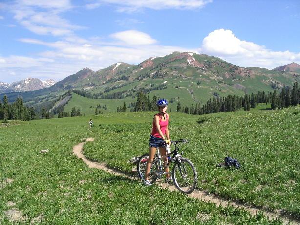 A woman in a red tank top and helmet stands next to a mountain bike on a grassy trail, surrounded by lush green mountains under a blue sky with fluffy white clouds. Trail 401 mountain bike trail.
