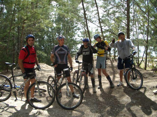 Group of five mountain bikers posing together on a dirt trail surrounded by trees. They are wearing helmets and biking gear, with their bicycles beside them. The scene captures a sense of camaraderie and adventure in an outdoor setting. Amelia Earhart Park mountain bike trail.