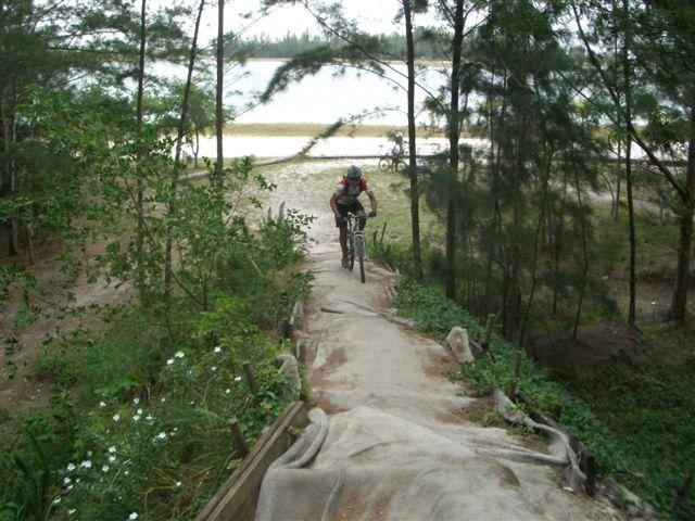 A cyclist riding down a dirt ramp, surrounded by trees and greenery, with a body of water visible in the background. Another cyclist can be seen in the distance on the trail. Amelia Earhart Park mountain bike trail.