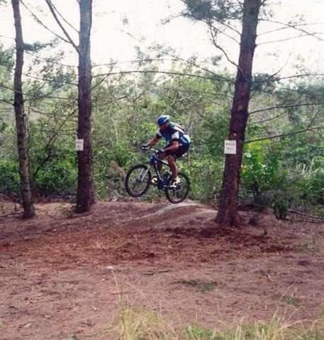 A mountain biker in a blue helmet and jersey leaps off a small hill among trees in a forested area. The ground is covered in dirt and pine needles, and signs are visible on the trees nearby. Amelia Earhart Park mountain bike trail.