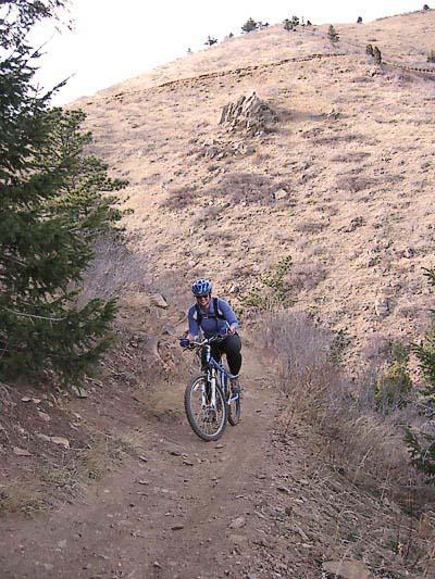 A mountain biker climbs a dirt trail on a hillside, surrounded by sparse vegetation and rocky terrain. The cyclist is wearing a blue helmet and a blue jacket, focused on navigating the path as the landscape slopes upward in the background. Chimney Gulch mountain bike trail.