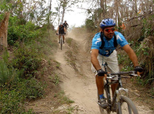 Two mountain bikers riding on a dirt trail through a forested area. The first rider is in the foreground, wearing a blue jersey and helmet, while the second rider follows behind, slightly obscured by dust. The surroundings include green vegetation and trees, suggesting an active outdoor environment. Amelia Earhart Park mountain bike trail.