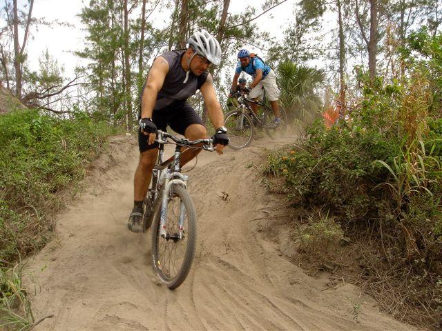 Two mountain bikers navigating a sandy trail in a forested area. The foreground biker, wearing a helmet and sporty attire, is leaning into a turn while kicking up dust, and a second biker can be seen in the background, also riding along the trail. The scene captures the dynamic nature of mountain biking amidst lush greenery and trees. Amelia Earhart Park mountain bike trail.