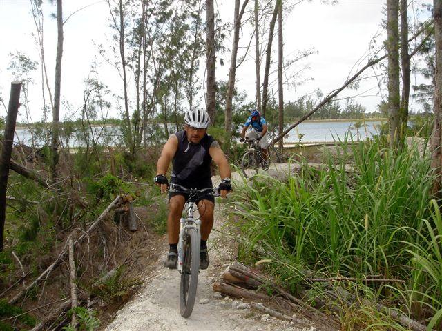 Two cyclists riding on a narrow dirt trail surrounded by trees and greenery. The first cyclist, in a sleeveless shirt and helmet, is in the foreground biking actively, while a second cyclist can be seen in the background near a body of water. The setting is natural and scenic, suggesting an outdoor adventure. Amelia Earhart Park mountain bike trail.