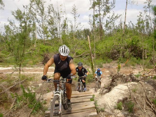 Three mountain bikers riding uphill on a wooden bridge in a wooded area. The landscape is green with trees in the background, and the sky is partly cloudy. One cyclist is leading the group, wearing a helmet and athletic gear. Amelia Earhart Park mountain bike trail.