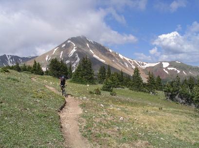 A mountain biker riding along a dirt trail surrounded by lush green grass and trees, with a backdrop of a majestic mountain peak partially covered in snow and a partly cloudy blue sky. Colorado Trail: Kenosha Pass To Breckenridge mountain bike trail.