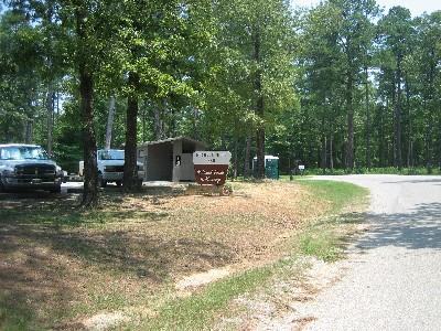 A gravel road curves around a wooded area, with several parked vehicles on the left. There is a small, rustic kiosk with a sign that reads "Welcome" visible near the road, surrounded by trees. A portable restroom is also present nearby. Noxubee Crest mountain bike trail.