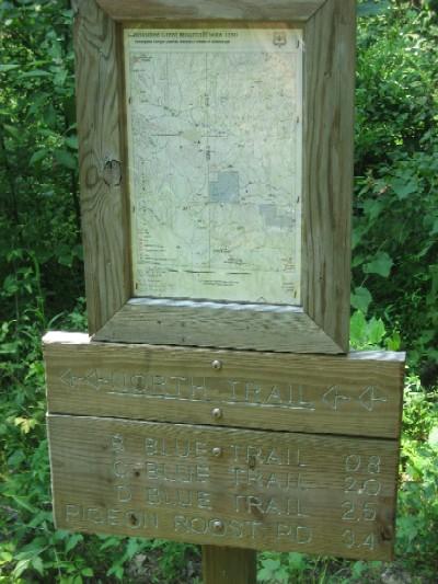 A wooden trail sign featuring a map at the top, indicating various hiking trails. The sign points towards the "North Trail" and lists distances to different routes: S Blue Trail (0.8 miles), C Blue Trail (2.0 miles), D Blue Trail (2.5 miles), and Pigeon Roost Road (3.4 miles). Surrounding vegetation is lush and green. Noxubee Crest mountain bike trail.