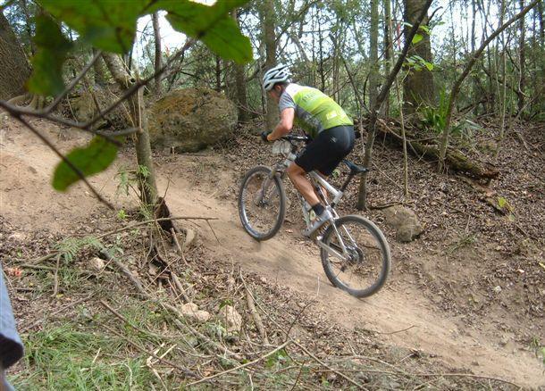 A mountain biker riding up a dirt trail surrounded by trees and foliage, showcasing an action shot in a natural setting. The cyclist is wearing a helmet and a green and white jersey, emphasizing the thrill of off-road biking. Santos mountain bike trail.