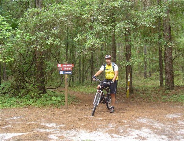 A person in cycling gear stands next to a bike at a trail intersection in a wooded area. The trail signs indicate directions for "John Brown" and "Ante Bellum" trails. The surroundings feature green foliage and pine trees, creating a natural outdoor setting. Santos mountain bike trail.