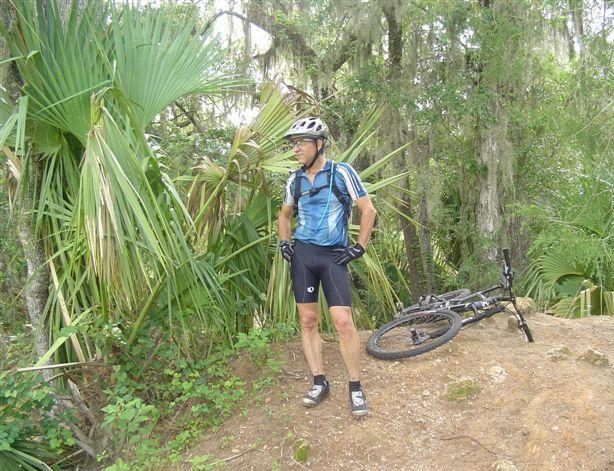 A cyclist in a blue and black jersey stands beside a mountain bike on a dirt path surrounded by lush green foliage. The cyclist is wearing a helmet, gloves, and padded shorts, looking towards the landscape. Santos mountain bike trail.