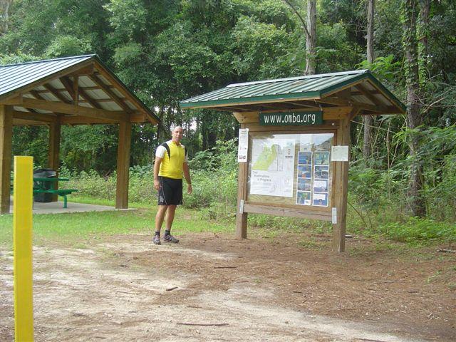 A person in a yellow shirt stands next to an informational map board at a wooded park, with a shelter structure visible in the background. The area is surrounded by greenery, indicating a natural setting for outdoor activities. Santos mountain bike trail.