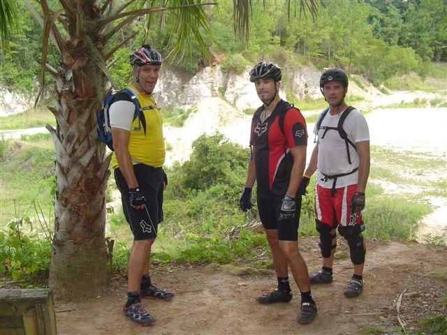 Three male mountain bikers wearing helmets and cycling gear pose for a photo on a dirt trail surrounded by greenery and palm trees. The background features a hilly landscape, suggesting an outdoor adventure setting. Santos mountain bike trail.