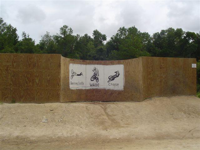 A wooden barrier with three illustrations and names painted on it. The images depict a person diving, a cyclist, and a motorcycle rider, with the names "Berrecloth," "WADE," and "Chase" accompanying each illustration. The setting is outdoors, surrounded by trees, and the ground appears to be dirt. Santos mountain bike trail.