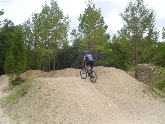 A mountain biker riding a dirt track with jumps, surrounded by lush green trees and a cloudy sky. The biker is positioned on a dirt mound, preparing to descend into a ramp. Santos mountain bike trail.
