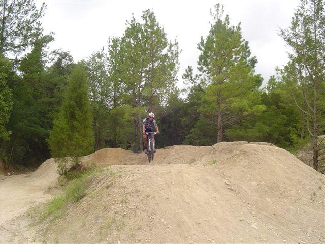 A mountain biker navigating a dirt trail with mounds in a forested area, surrounded by tall pine trees. The biker is mid-jump, showcasing an action shot in a natural outdoor setting. Santos mountain bike trail.