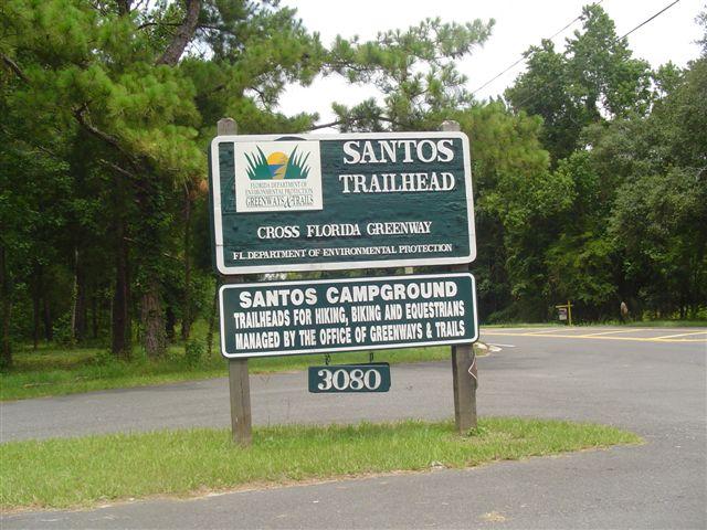A green and white sign indicating the Santos Trailhead, part of the Cross Florida Greenway, managed by the Florida Department of Environmental Protection. The sign displays information about the Santos Campground and trailheads for hiking, biking, and equestrian activities. The location number 3080 is also visible, set against a backdrop of trees and natural surroundings. Santos mountain bike trail.