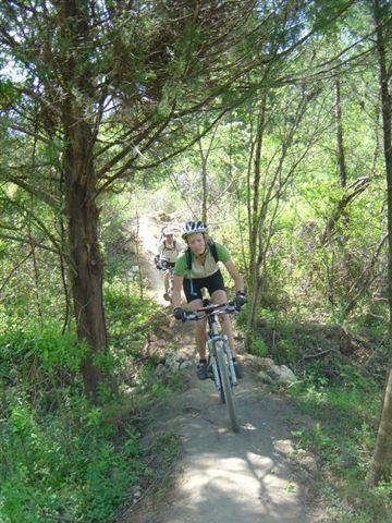 A mountain biker navigating a dirt trail surrounded by trees and greenery. The rider is wearing a helmet and an athletic outfit, focused on the path ahead. Another cyclist can be seen in the background, also riding on the trail. Santos mountain bike trail.