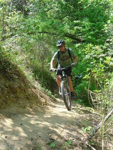 A person riding a mountain bike on a dirt trail surrounded by lush greenery and trees. The rider is wearing a helmet and is captured mid-motion, with one wheel lifted off the ground. Santos mountain bike trail.