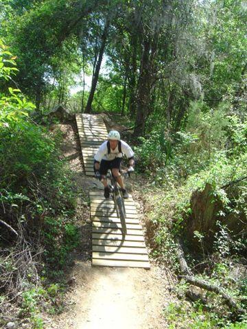 A mountain biker descending on a wooden bridge over a trail in a lush, green forest. The cyclist is wearing a helmet and riding through a natural setting with dense foliage and trees surrounding the path. Santos mountain bike trail.