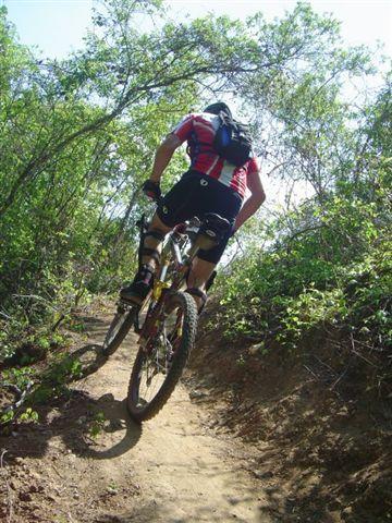 A cyclist in a red and black jersey rides uphill on a narrow dirt path surrounded by lush greenery and bushes. The image captures the back view of the cyclist, highlighting the bike's tires against the earthy trail. Santos mountain bike trail.