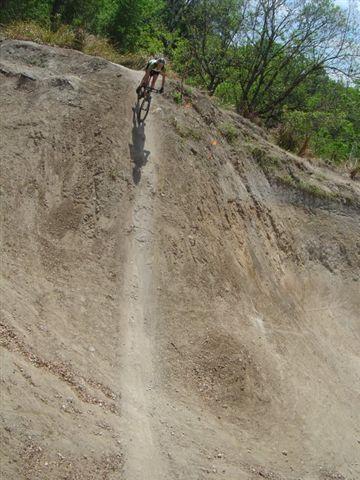 A cyclist riding up a steep, dusty slope surrounded by greenery. The trail shows signs of use, with a clear path carved into the dirt. Santos mountain bike trail.