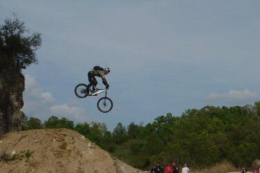 A cyclist performing a jump on a BMX bike over a dirt ramp, with spectators visible in the background and a green landscape under a blue sky. Santos mountain bike trail.