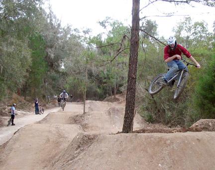 A mountain biker in a red shirt performs a jump over a dirt jump trail in a forested area, with another biker in the background and spectators watching nearby. Santos mountain bike trail.
