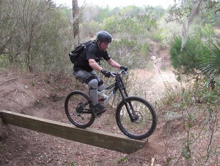 A mountain biker in safety gear rides over a narrow wooden bridge on a dirt trail surrounded by greenery. Santos mountain bike trail.