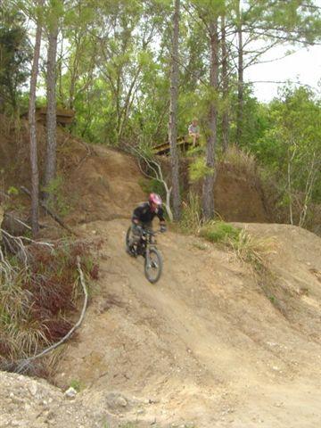 A person wearing a helmet and black clothing rides a mountain bike down a dirt slope in a wooded area. Trees and shrubs surround the scene, and a figure can be seen in the background on a wooden structure. Santos mountain bike trail.