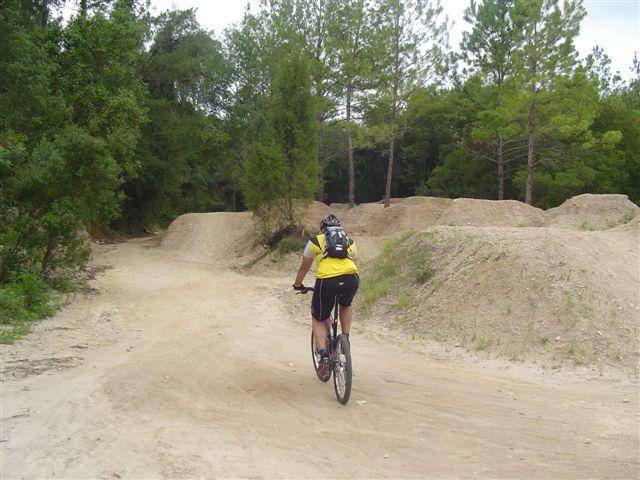 A person riding a mountain bike on a dirt trail surrounded by trees, with dirt jumps visible in the background. The cyclist is wearing a yellow shirt and a helmet, and the trail is winding through a natural, wooded area. Santos mountain bike trail.