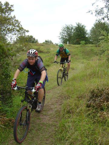 Two mountain bikers navigate a dirt trail through a grassy area, surrounded by trees. One rider is in the foreground, wearing a helmet and gloves, while the other follows closely behind. The scene captures the essence of outdoor biking and adventure. Balm Boyette Scrub Preserve mountain bike trail.