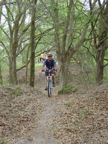 A person riding a mountain bike along a narrow trail surrounded by trees and greenery. The trail is lined with fallen leaves, and a few other cyclists can be seen in the background. Balm Boyette Scrub Preserve mountain bike trail.