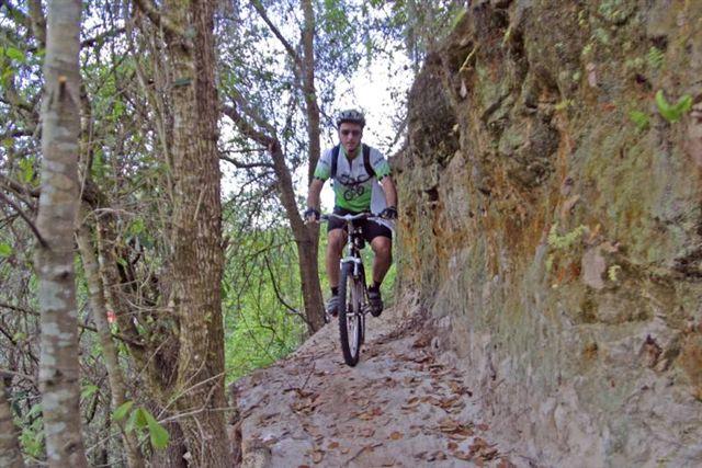 A cyclist navigating a narrow, rocky trail surrounded by trees, wearing a helmet and a backpack. The path is flanked by a steep rock face and greenery, indicating a scenic outdoor environment. Balm Boyette Scrub Preserve mountain bike trail.