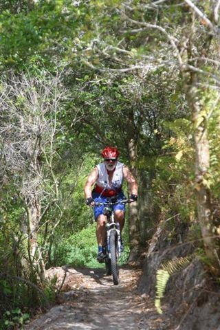 A person riding a mountain bike along a narrow trail surrounded by greenery and trees, wearing a red helmet and a sleeveless shirt. The scene captures a moment of outdoor adventure in a natural setting. Balm Boyette Scrub Preserve mountain bike trail.