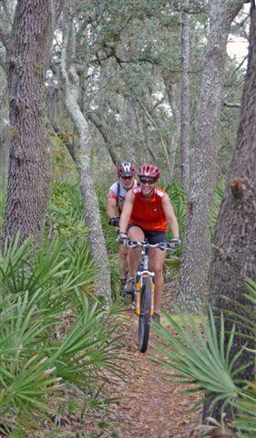 Two mountain bikers riding on a narrow trail through a forest, surrounded by trees and lush green vegetation. One rider is in the foreground wearing a red shirt and helmet, while the other is following closely behind. Balm Boyette Scrub Preserve mountain bike trail.