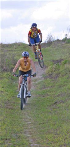 Two cyclists navigate a grassy trail. The cyclist in the foreground is riding downhill on a mountain bike, while a second cyclist in a colorful jersey is airborne, performing a jump off a small ridge. The scene is set against a cloudy sky, with greenery surrounding the trail. Balm Boyette Scrub Preserve mountain bike trail.