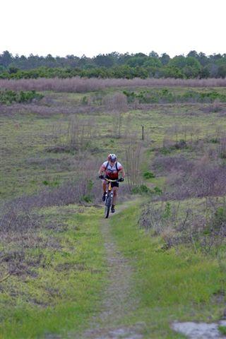 A cyclist riding a mountain bike on a narrow dirt trail through a grassy landscape with sparse vegetation and distant trees under a cloudy sky. Balm Boyette Scrub Preserve mountain bike trail.