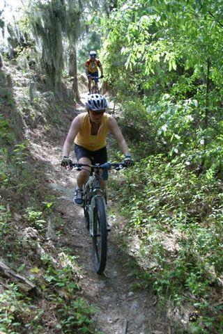 A woman rides a mountain bike on a narrow, lush green trail surrounded by trees and undergrowth. Another cyclist can be seen in the background, also navigating the trail. The scene captures a sunny day in a natural setting. Balm Boyette Scrub Preserve mountain bike trail.