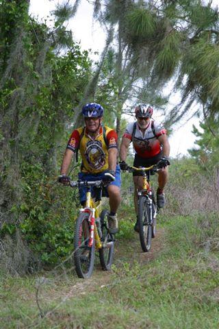 Two cyclists riding mountain bikes along a narrow dirt trail surrounded by green foliage. One cyclist, in a blue and yellow jersey, leads the way, while the other follows closely behind in a red and black jersey. The scene captures a moment of outdoor adventure in a natural setting. Balm Boyette Scrub Preserve mountain bike trail.
