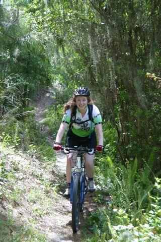 A woman riding a mountain bike on a narrow trail surrounded by lush greenery and trees. She is wearing a helmet and a cycling jersey, smiling as she navigates the path. Balm Boyette Scrub Preserve mountain bike trail.