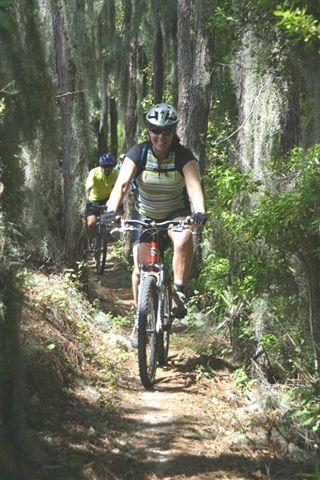 Two mountain bikers riding along a narrow dirt trail surrounded by lush greenery and tall trees. The foreground features one rider wearing a helmet and a striped shirt, smiling as they navigate the path, while another rider is slightly behind, partially obscured by the foliage. Balm Boyette Scrub Preserve mountain bike trail.