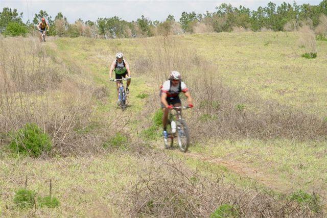 Three mountain bikers ride along a grassy trail in a natural setting. The foreground features two riders navigating the uneven terrain, while a third rider can be seen further in the background. The area is surrounded by sparse vegetation and distant trees under a cloudy sky. Balm Boyette Scrub Preserve mountain bike trail.